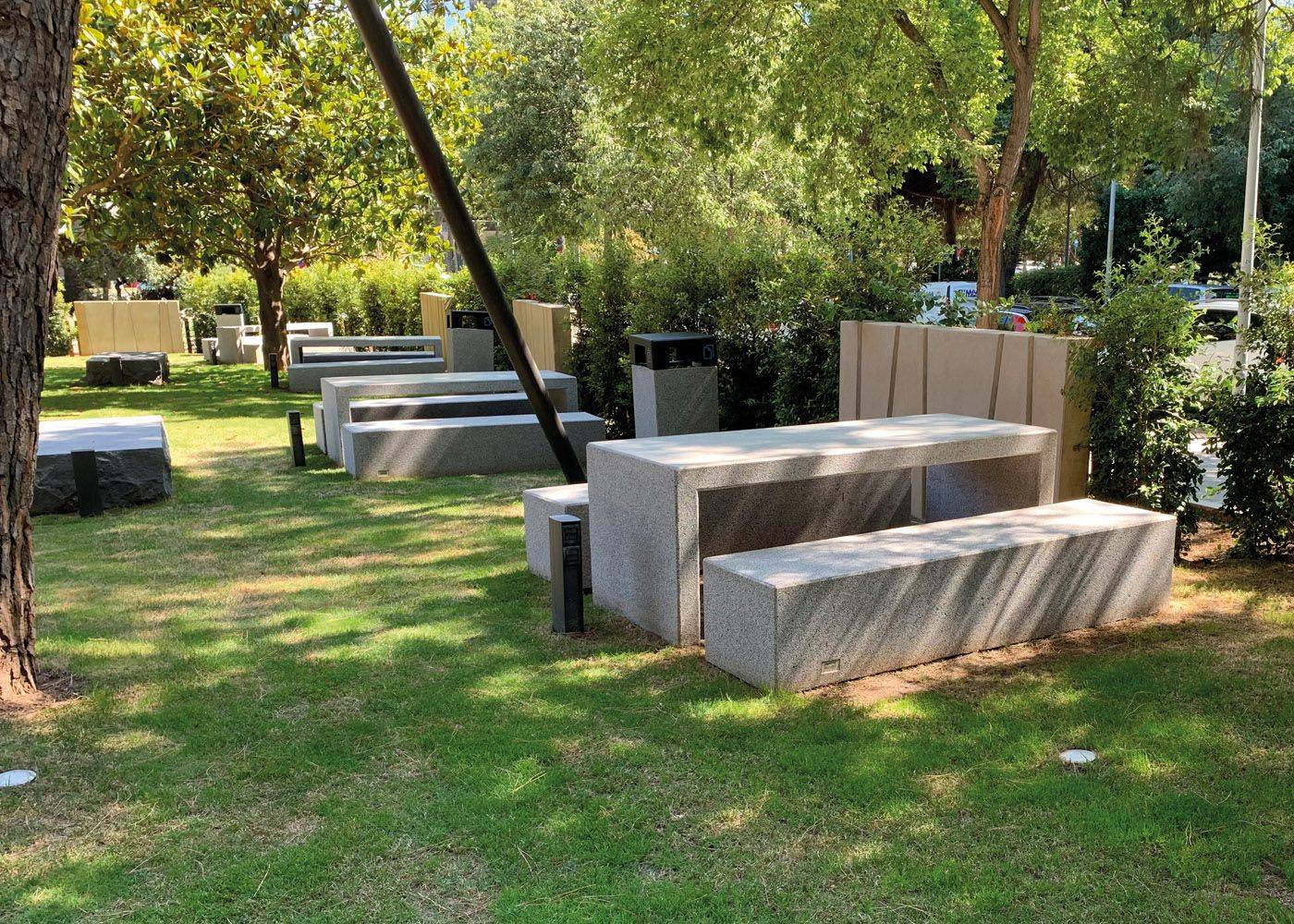 Vista de mesas y bancos de hormigón gris en un espacio verde, ideal para áreas recreativas y jardines públicos.