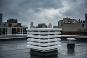 Aspirador estatico ortogonal de hormigon color blanco con laminas horizontales instalado sobre la salida de humos del tejado plano de un atico urbano bajo lluvia ligera, edificios de fondo desenfocados, ambiente nublado