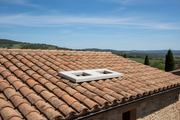 Base doble de hormigon color gris ciment con dos aberturas rectangulares instalada sobre el tejado de tejas de una masia rural de piedra, cielo azul con nubes suaves