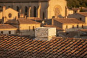 Remate de chimenea de piedra color champagne instalado sobre el tejado de teja plana de un edificio historico de casco antiguo, fachadas de piedra desenfocadas al fondo, luz calida de atardecer
