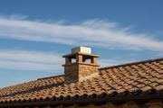 Remate de chimenea de piedra color champagne instalado sobre el tejado de una masia rural, cielo azul con nubes suaves