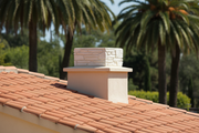 Remate de chimenea de piedra color Pirineos instalado sobre el tejado de teja curva de una villa mediterranea blanca, jardin con palmeras desenfocado al fondo, luz de mediodia
