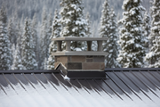 Sombrero de chimenea de hormigon color gris con patas y goterones instalado sobre la salida de humos del tejado metalico oscuro nevado de una cabana de montana, pinos nevados desenfocados al fondo, luz de invierno