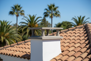 Sombrero de chimenea de hormigon color gris claro con patas y goterones instalado sobre la salida de humos del tejado de teja curva de una villa mediterranea blanca, jardin con palmeras desenfocado al fondo, luz de mediodia